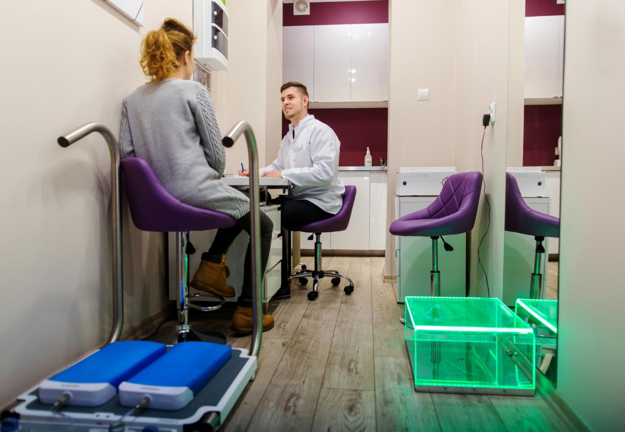 Konsultacja dietetyczna: Klientka siedzi na fioletowym krześle obok urządzenia do analizy stóp, dietetyk w białym fartuchu notuje przy biurku.