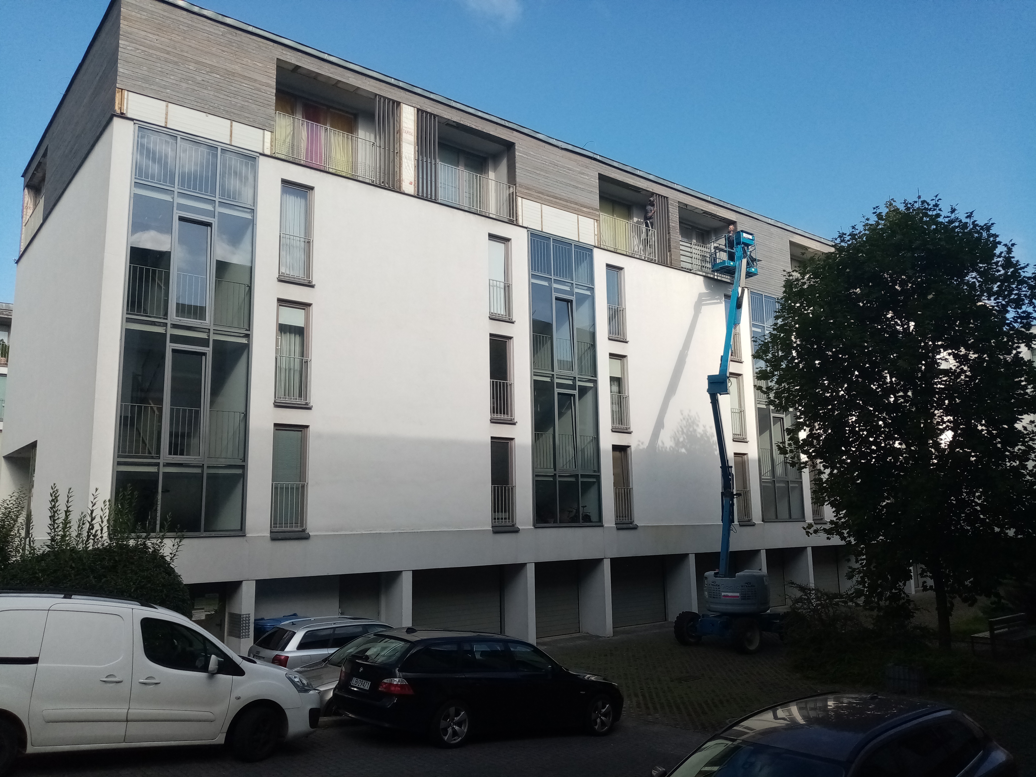 Pracownik na podnośniku koszowym wykonuje prace na elewacji budynku wielorodzinnego z drewnianym wykończeniem, widoczne zaparkowane samochody i zieleń.