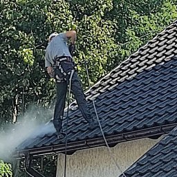 Alpin Group Marcin Bedryjczuk i Mirosław Wierzbicki - Pracownik w uprzęży zabezpieczającej czyści dach z czarnej dachówki za pomocą myjki ciśnieniowej, widoczny strumień wody i drabina oparta o ścianę budynku.