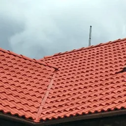 Czerwony dach z dachówki falistej z oknem dachowym i anteną na tle pochmurnego nieba.