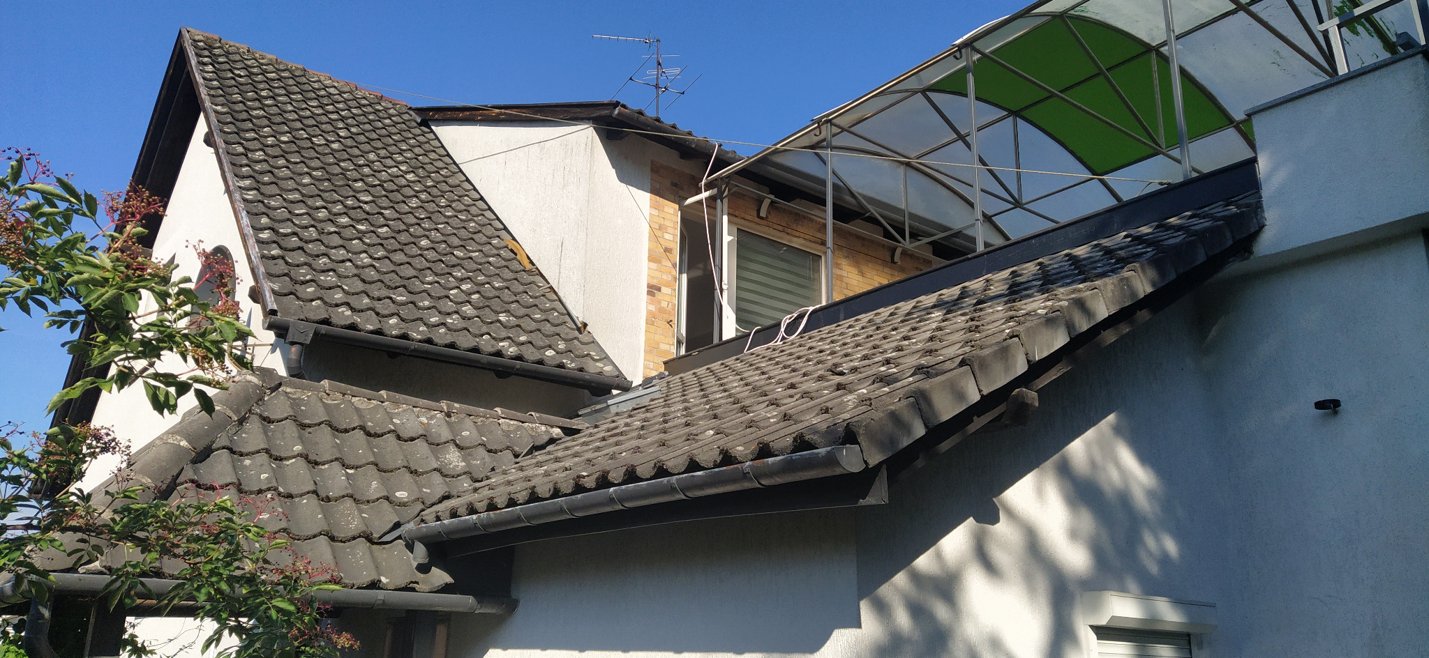 Fragment dachu z szarymi dachówkami, widoczne rynny, fragment ściany budynku i zadaszenie nad oknem, w tle antena telewizyjna.
