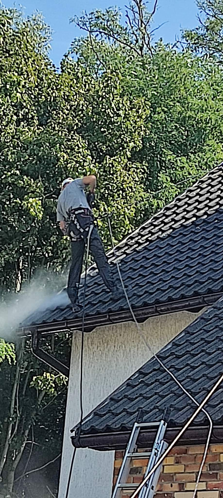 Pracownik w uprzęży zabezpieczającej czyści dach z czarnej dachówki za pomocą myjki ciśnieniowej, widoczny strumień wody i drabina oparta o ścianę budynku.