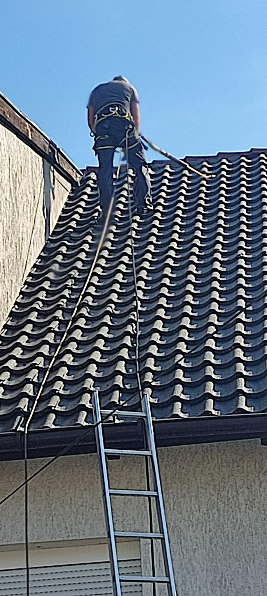 Pracownik w uprzęży zabezpieczającej stoi na dachu z ciemnej dachówki ceramicznej, widoczna drabina oparta o ścianę budynku, niebieskie niebo w tle.
