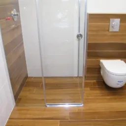 Nowoczesna łazienka z kabiną prysznicową i podwieszaną toaletą. Płytki imitujące drewno na podłodze i częściowo na ścianach, białe płytki na pozostałej powierzchni.