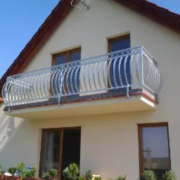 Kuty, metalowy balkon z dekoracyjnymi elementami na tle jasnej elewacji domu jednorodzinnego. Widok z dołu, słoneczny dzień.