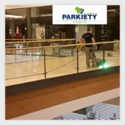 Pracownik w ciemnej koszulce z logo firmy obsługuje maszynę do renowacji drewnianej podłogi w przestrzeni handlowej, widoczne szklane barierki i sklepy w tle.