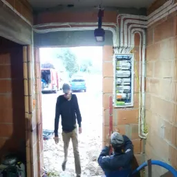 Wnętrze w trakcie prac elektrycznych: widoczna rozdzielnica, kable prowadzone po ścianach z cegły, dwóch mężczyzn w trakcie pracy, jeden z poziomicą, drugi wchodzący do pomieszczenia.