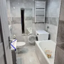 Wyremontowana łazienka w odcieniach szarości z pralką, grzejnikiem drabinkowym i wiadrem grzybów w wannie.