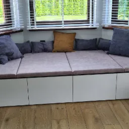 Wnękowe siedzisko z szufladami, obite jasnoróżową tkaniną, z poduszkami w odcieniach szarości i brązu, umieszczone pod oknem z żaluzjami.