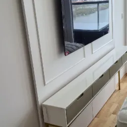 Biała szafka RTV z szarymi szufladami i złotymi nogami, wbudowana w ścianę z dekoracyjnymi listwami, z telewizorem wiszącym nad nią, odbijającym widok miasta.