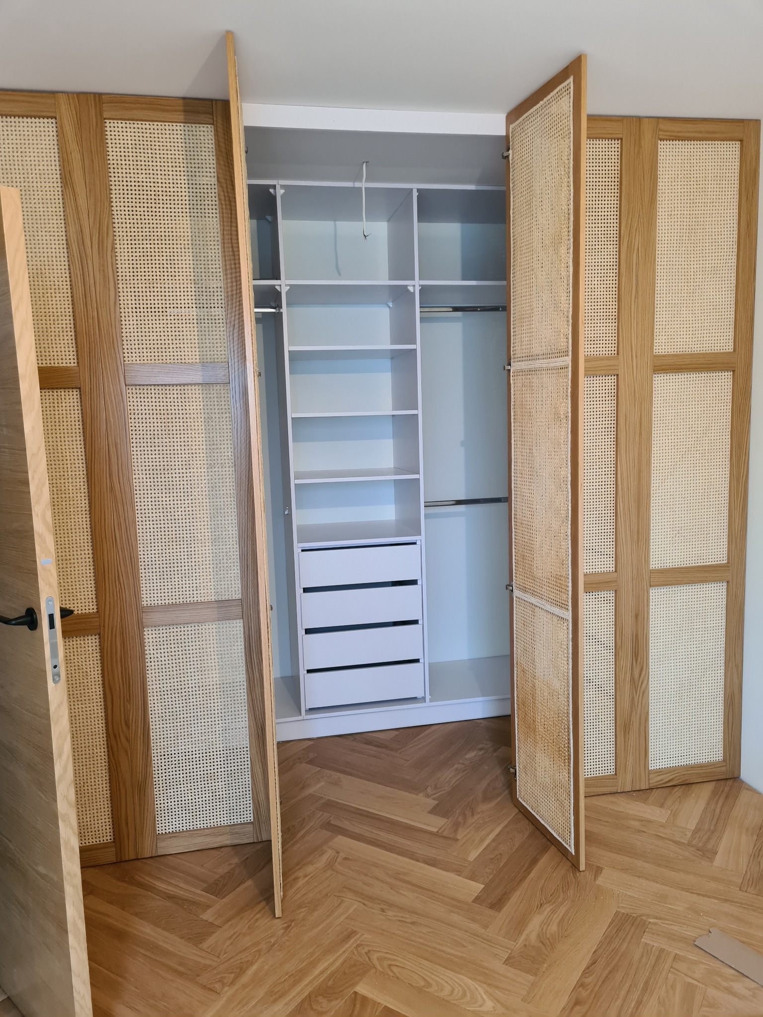 Otwarta szafa wnękowa z drewnianymi drzwiami wypełnionymi plecionką rattanową, ukazująca białe wnętrze z półkami i szufladami, na podłodze parkiet w jodełkę.