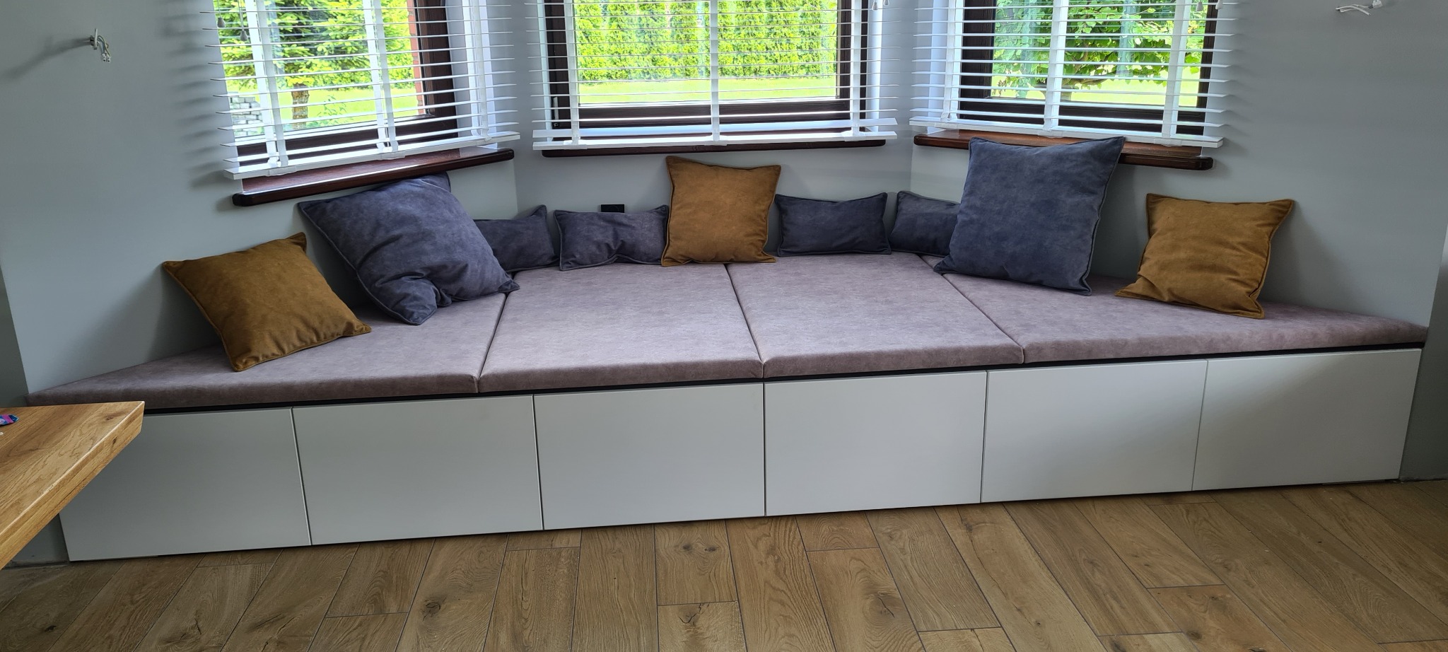 Wnękowe siedzisko z szufladami, obite jasnoróżową tkaniną, z poduszkami w odcieniach szarości i brązu, umieszczone pod oknem z żaluzjami.