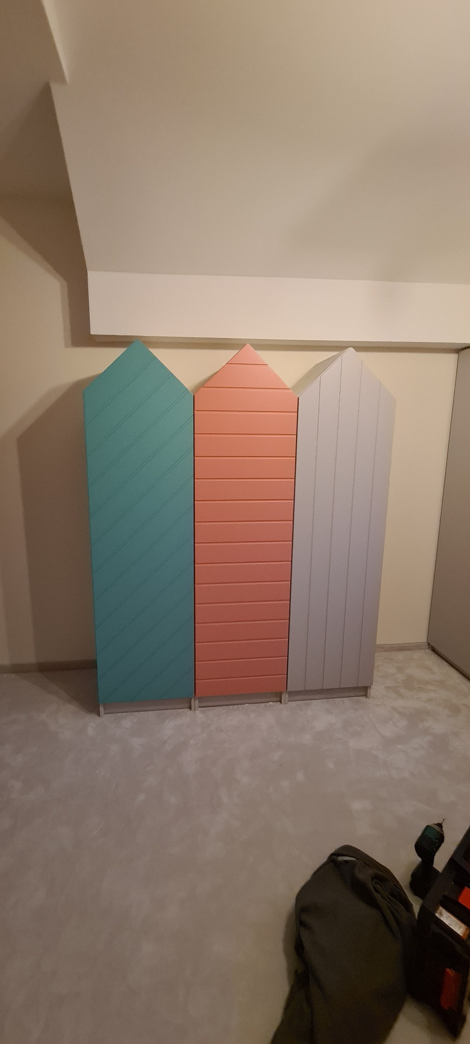 Trzy kolorowe, niskie szafy z drewnianych paneli w kształcie domków dla dzieci, ustawione obok siebie na szarym dywanie, w tle narzędzia monterskie.