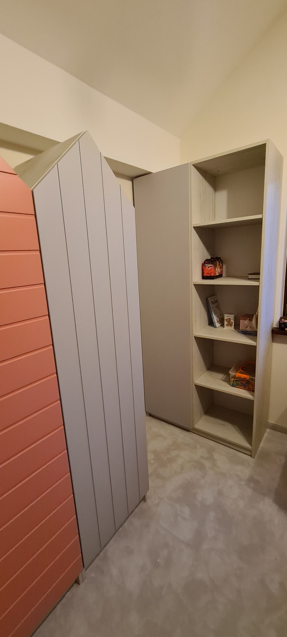 Narożna szafa z jasnoszarego drewna z półkami, na których stoją przedmioty dekoracyjne i książki, widoczna obok szafa z drzwiami w kształcie domku pomalowanymi na jasny róż i szaro.