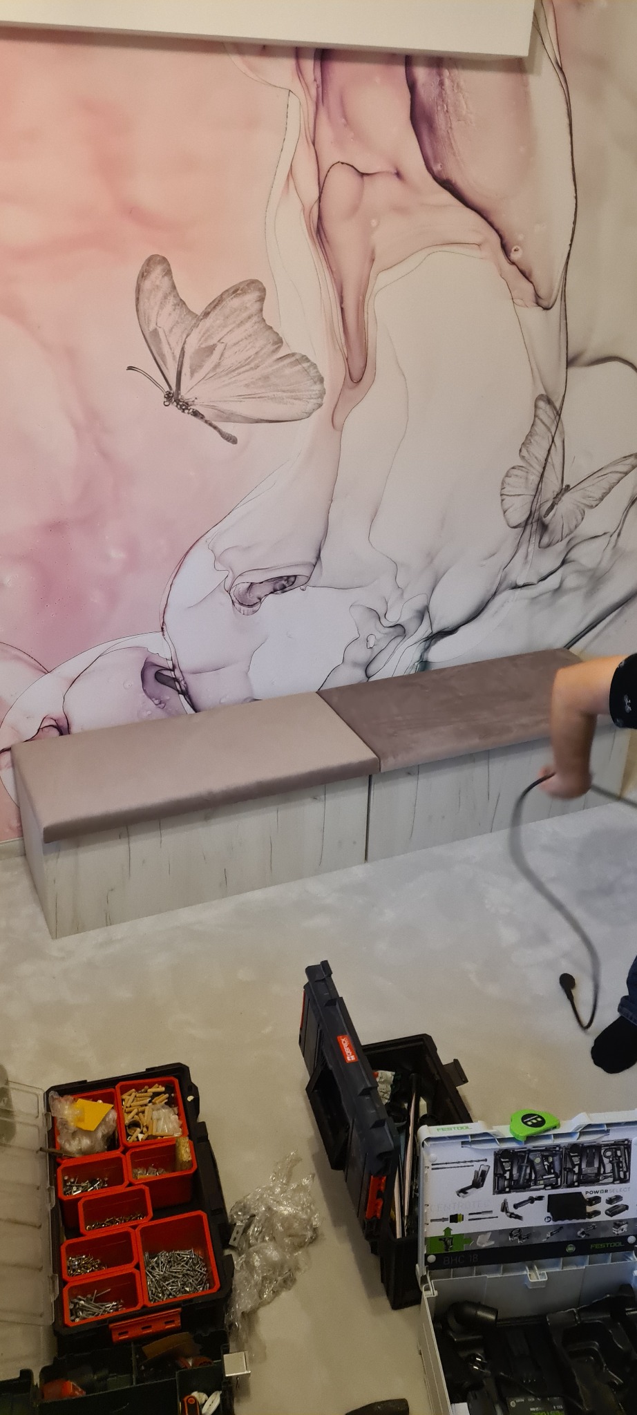 Montaż siedziska tapicerowanego na tle ściany z motywem motyli, widoczne skrzynki z narzędziami i fragment dłoni montera.