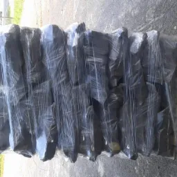 Paleta z czarnym brykietem węglowym, owinięta folią stretch, widoczna tekstura brykietu i nierówności folii, tło z zielenią drzew.