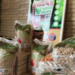 Stos worków z pelletem drzewnym, widoczne oznaczenia jakości EN A1 plus i logo Tartak Olczyk, w tle paleta z workami i tablica reklamowa z informacjami kontaktowymi.