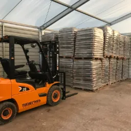 Pomarańczowy wózek widłowy marki Hangcha z butlą gazową, w magazynie z metalowymi ścianami, obok palet z workami z nieznaną zawartością owiniętymi folią stretch.