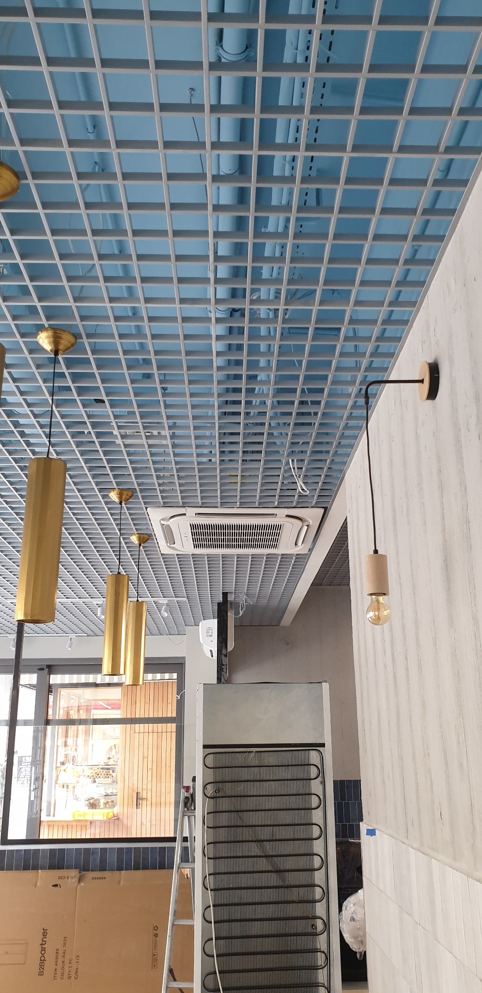 Kasetonowy klimatyzator GREE wkomponowany w sufit podwieszany z kratką, widoczne rury instalacyjne, designerskie lampy wiszące i kinkiet, chłodziarka w tle.