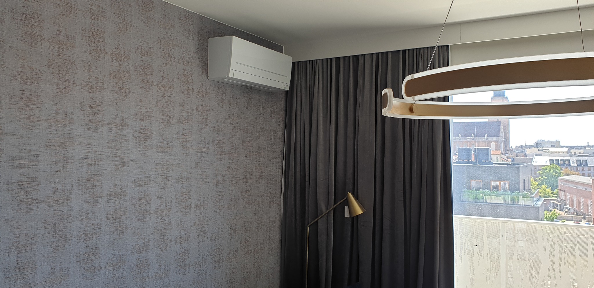Biały klimatyzator Mitsubishi Electric MSZ-AP zamontowany na ścianie z szarą tapetą w pokoju z szarymi zasłonami, złotą lampą podłogową i widokiem na miasto.