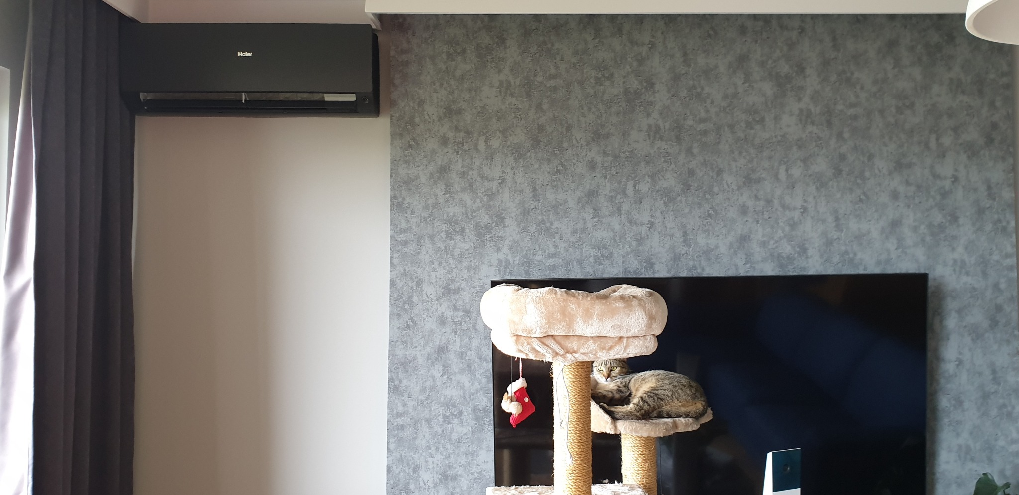 Czarny matowy klimatyzator HAIER FLEXIS zamontowany na ścianie w salonie, obok szarych zasłon i ściany o teksturze betonu; na pierwszym planie drapak dla kota z leżącym kotem.