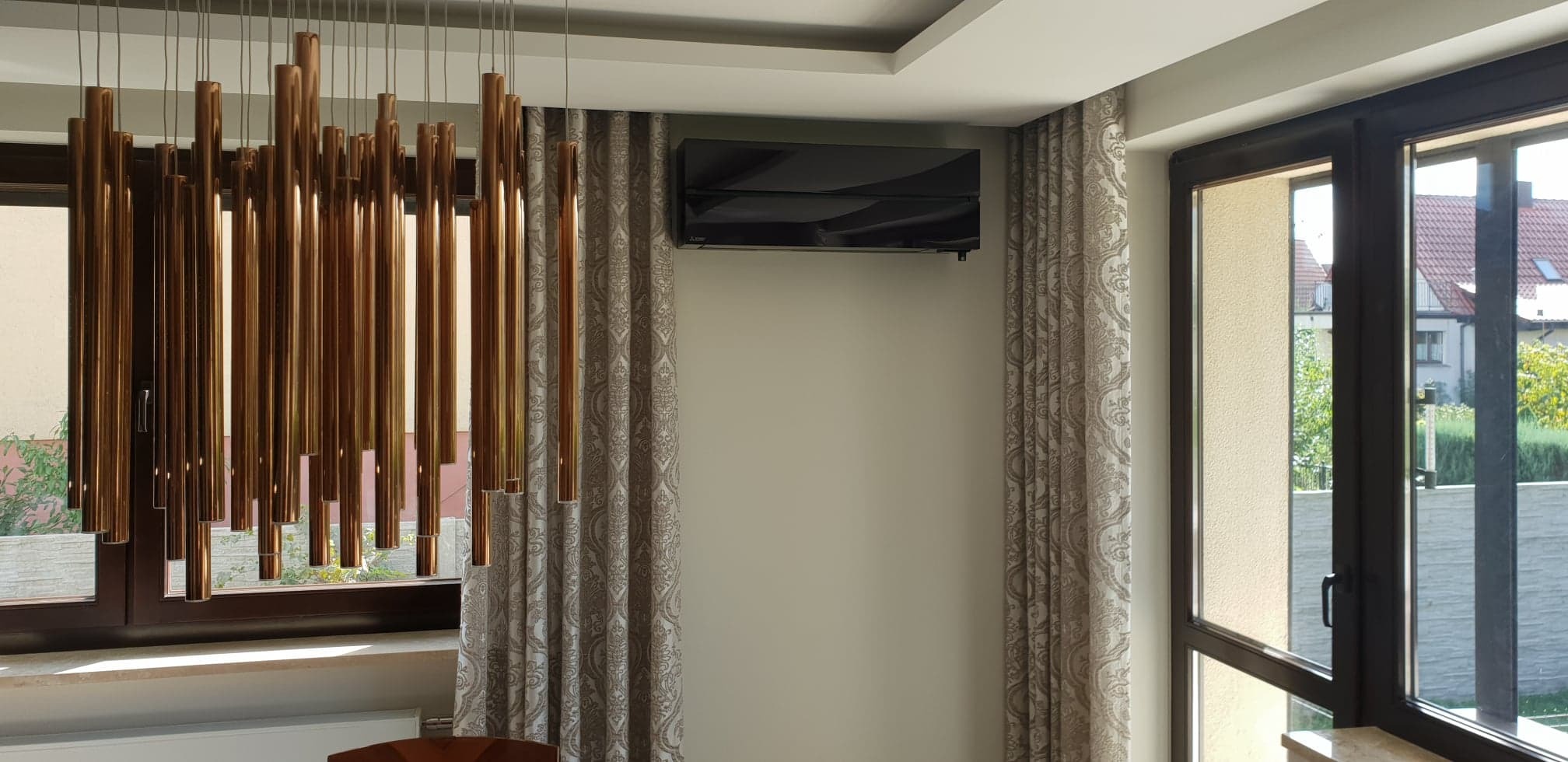 Czarny klimatyzator Mitsubishi Electric serii MSZ-LN zamontowany na ścianie w nowoczesnym salonie, widoczne zasłony w ornamenty i dekoracyjna lampa z miedzianych rurek.