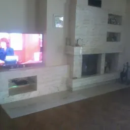 Wykończony kominek z jasnego kamienia w salonie z drewnianą podłogą. Na ścianie telewizor i dekoracyjne półki. Elementy wykończenia dopasowane kolorystycznie.