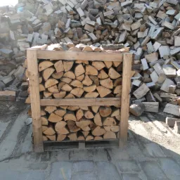 Drewno kominkowe w drewnianej skrzyni na palecie, w tle sterta drewna. Jasne oświetlenie podkreśla strukturę drewna. Gotowy produkt do transportu.