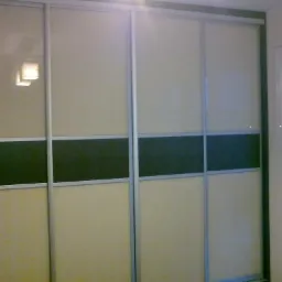 Czterodrzwiowa szafa przesuwna z beżowymi i ciemnymi panelami, obramowana srebrnymi profilami, z widocznym odbiciem lampy i fragmentem okna z żaluzjami.