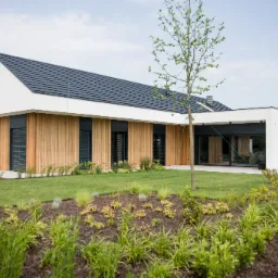 Nowoczesny dom jednorodzinny z drewnianą elewacją i ciemną dachówką, otoczony zadbanym trawnikiem i rabatami z roślinami ozdobnymi.