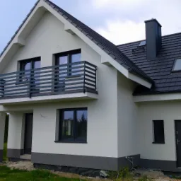 Nowoczesny dom jednorodzinny z białą elewacją, ciemną stolarką okienną i balkonem z metalową balustradą, dach pokryty czarną dachówką, widoczne ślady prac wykończeniowych wokół budynku.