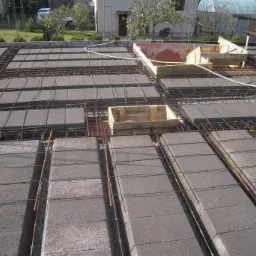 Wieniec stropowy z szalunkami i zbrojeniem, widok z góry. Elementy betonowe ułożone między żebrami konstrukcji. W tle budynki mieszkalne i zieleń.