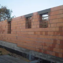 Ściana domu w trakcie budowy z czerwonej cegły ceramicznej, widoczne otwory okienne i betonowy fundament. Częściowo zarośnięty teren wokół budynku.