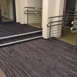 Korytarz z czarno-białą wykładziną w prążki, zabezpieczony barierką i pachołkiem ostrzegawczym przy schodach.