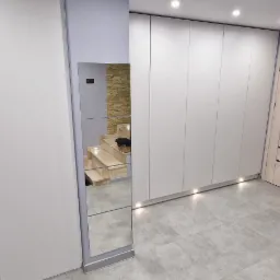 Wnętrze przedpokoju z białą szafą na wymiar, lustrem odbijającym schody z kamienną okładziną oraz siedziskiem z panelem tapicerowanym w kolorze czarnym.