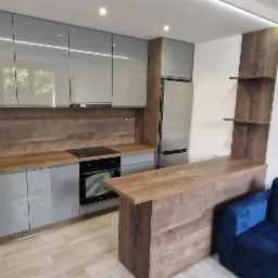 Nowoczesna kuchnia z szarymi, lakierowanymi szafkami, blatem w kolorze drewna i barkiem, uzupełniona o niebieską sofę w tle.