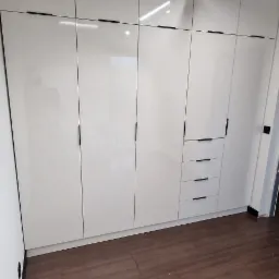 Biała zabudowa meblowa od podłogi do sufitu z minimalistycznymi, czarnymi uchwytami, uzupełniona o szuflady, na tle brązowej podłogi imitującej drewno.