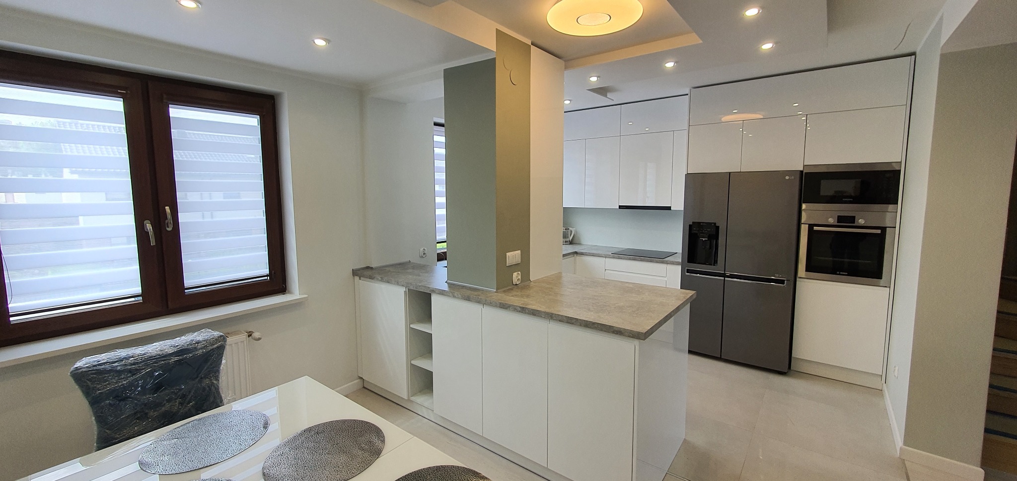 Nowoczesna, jasna kuchnia z białymi szafkami, szarym blatem i sprzętem AGD w zabudowie. Widoczny półwysep kuchenny z otwartymi półkami i szarym blatem. Krzesło owinięte folią ochronną przy stole.