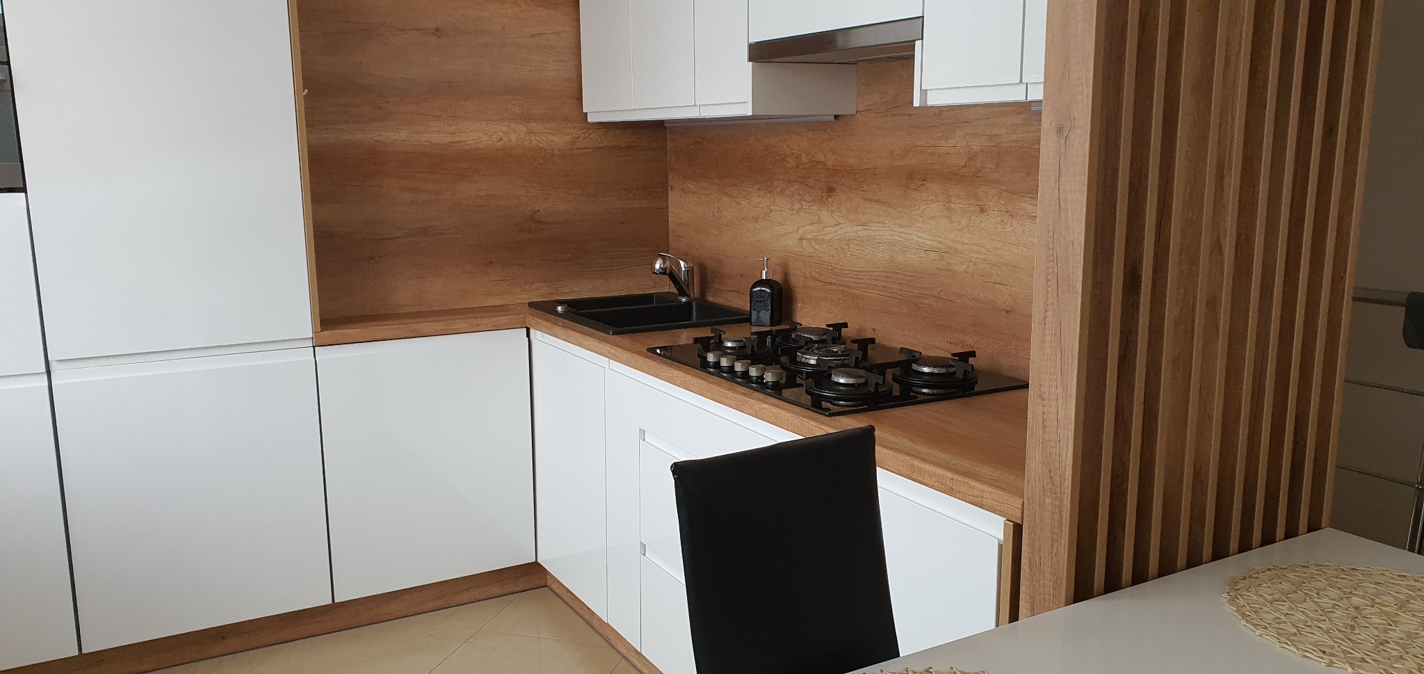 Nowoczesna kuchnia: białe szafki, drewnopodobny blat i panel ścienny z pionowych listew, czarny zlew i płyta grzewcza, widoczny fragment krzesła.