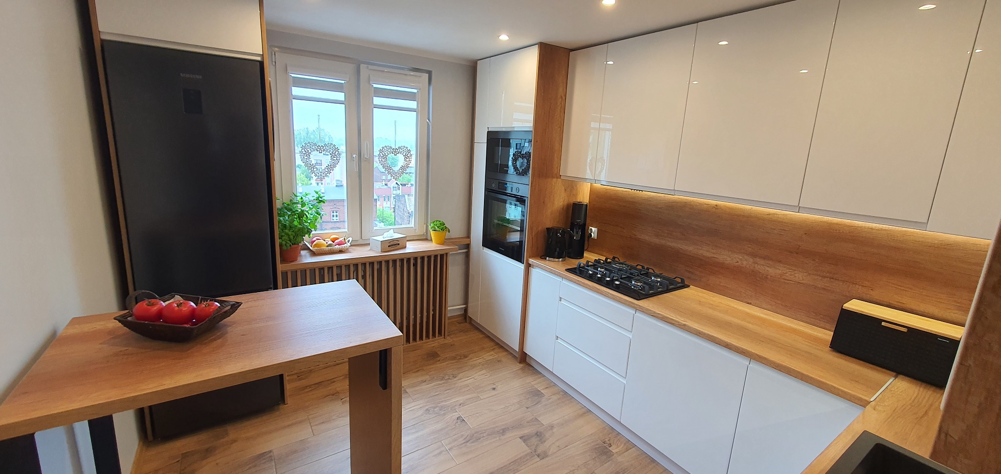 Nowoczesna kuchnia: białe, lakierowane szafki z drewnianymi blatami i pasem nad blatem, czarny piekarnik i płyta gazowa, drewniany stół z owocami, okno z dekoracjami w kształcie serc.