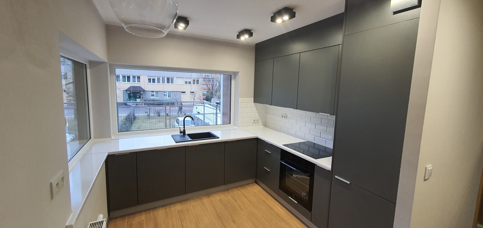 Nowoczesna kuchnia z szarymi szafkami, białym blatem i czarnym zlewem pod oknem z widokiem na budynek mieszkalny.