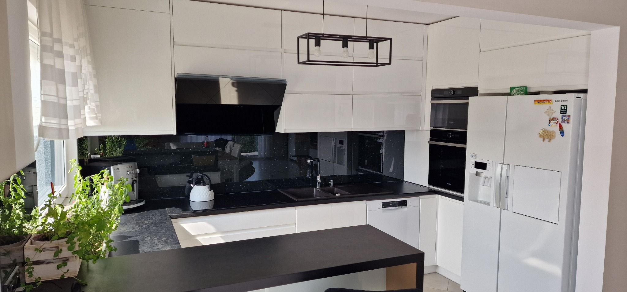 Nowoczesna kuchnia z białymi, lakierowanymi frontami, czarnym blatem z brokatowym wzorem, szklanym panelem nad blatem, wiszącą lampą w stylu industrialnym i roślinami w oknie.