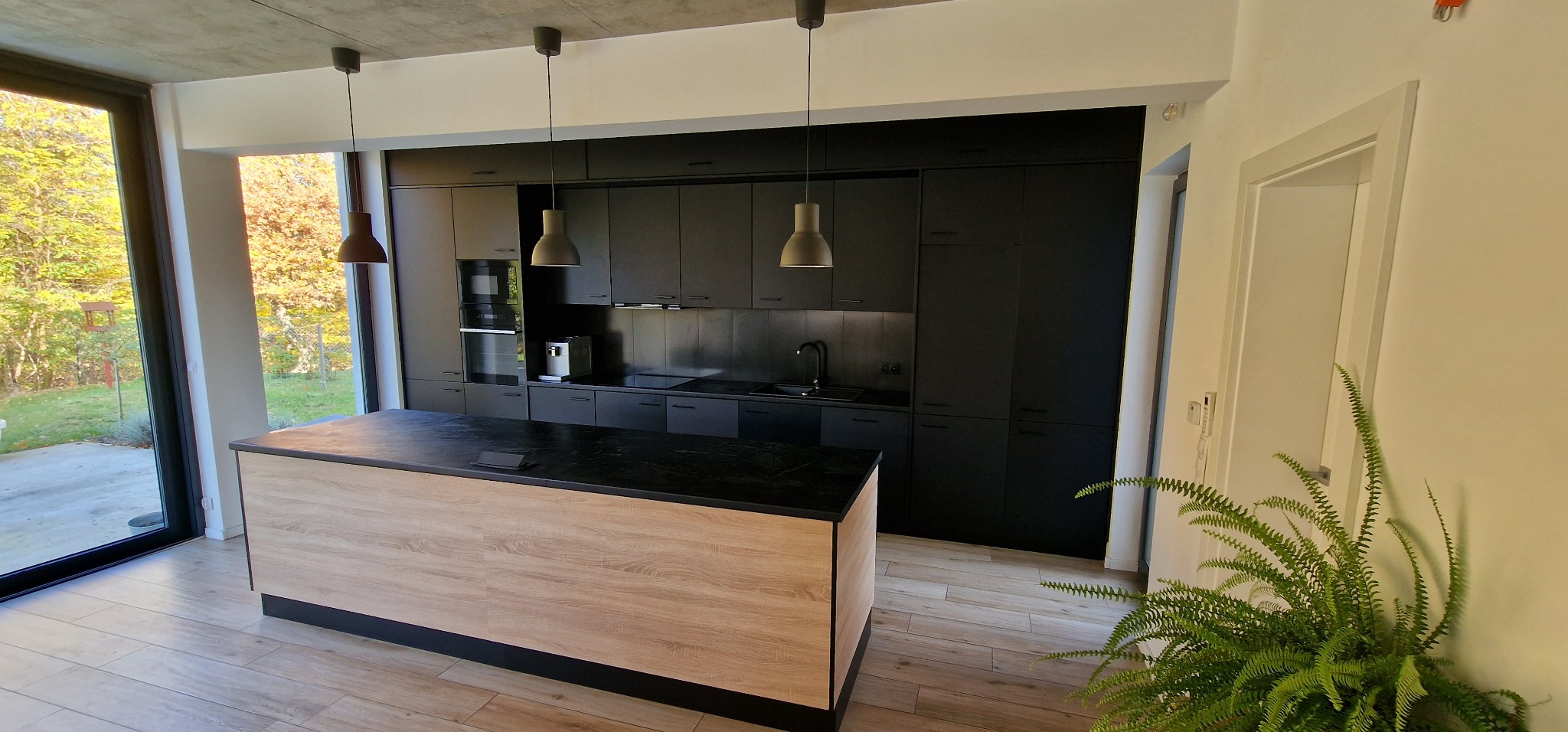 Nowoczesna kuchnia z czarnymi szafkami i wyspą z blatem z ciemnego kamienia oraz drewnopodobną okleiną, oświetlona trzema wiszącymi lampami, z widokiem na zieleń za oknem.