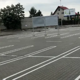 Nowo wybrukowany parking z szarej kostki brukowej z wyznaczonymi białymi liniami miejsc parkingowych, widoczne dwa puste bilbordy reklamowe w tle.