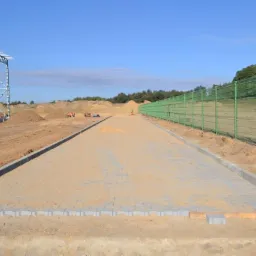 Trwa budowa drogi z kostki brukowej; widoczne ułożone fragmenty nawierzchni, obrzeża betonowe, przygotowane podłoże, a w tle stosy ziemi i konstrukcja stalowa. Na drugim planie pracownicy...