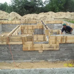 Budowa fundamentów domu z bloczków betonowych, z drewnianym szalunkiem i zbrojeniami. Pracownik w zielonej czapce przy pracy. Data budowy w prawym dolnym rogu.