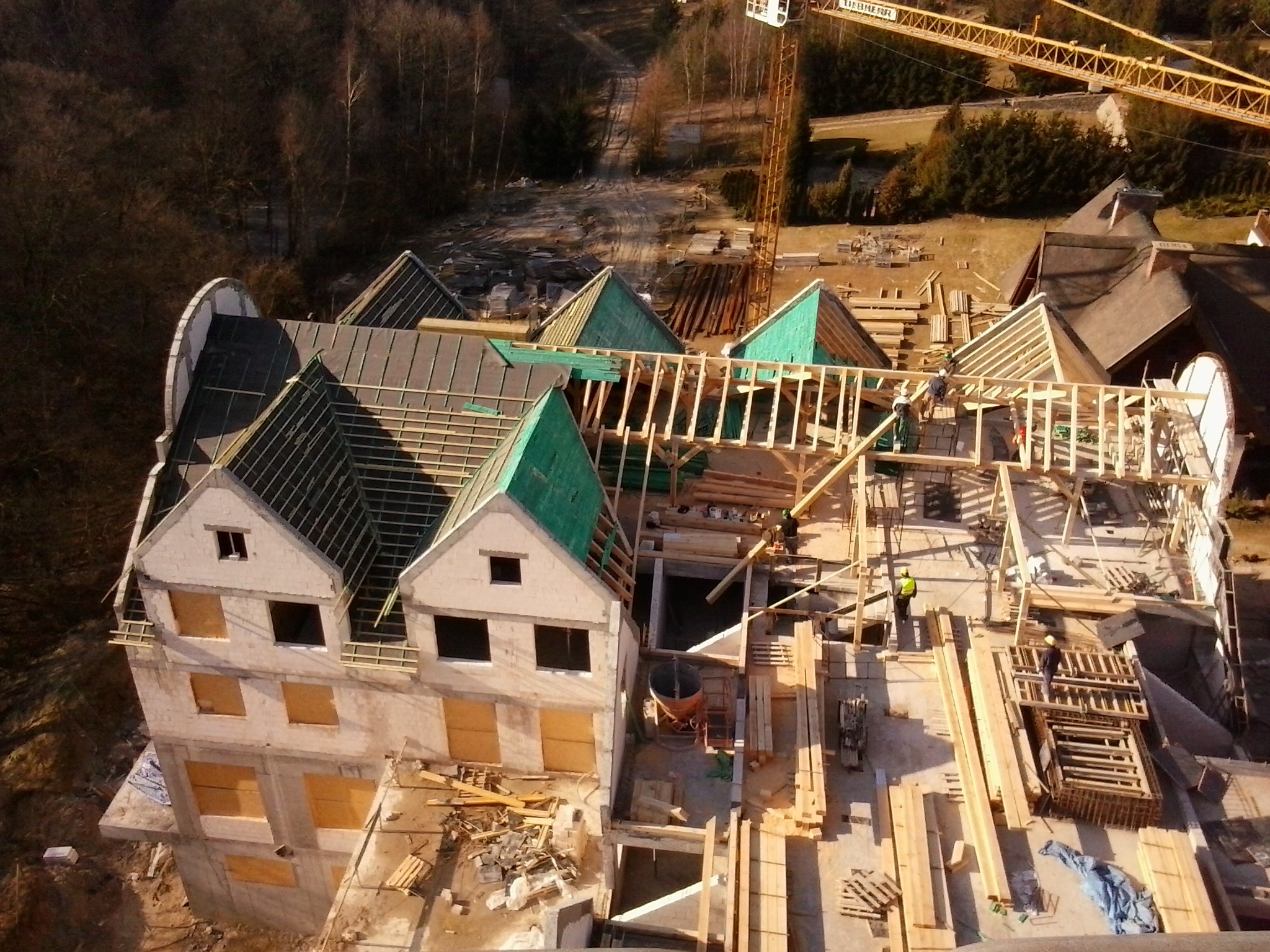 Wysoki kąt widzenia na budowę domu z widoczną konstrukcją dachu, żółtym dźwigiem w tle i pracownikami w kaskach.