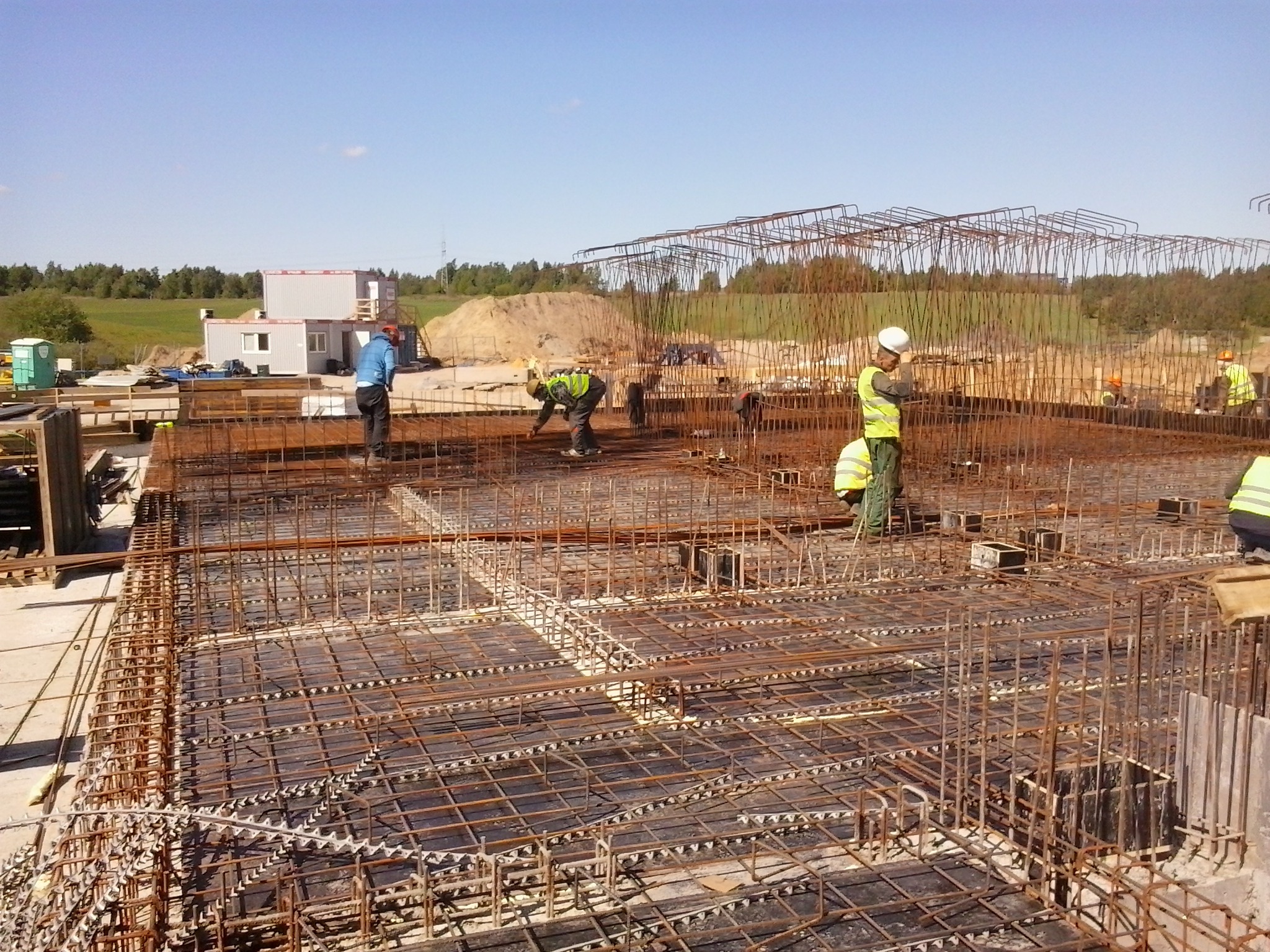 Prace zbrojarskie na budowie: widok na konstrukcję z siatki stalowej, robotnicy w odblaskowych kamizelkach i kaskach, w tle barakowóz i hałda ziemi.