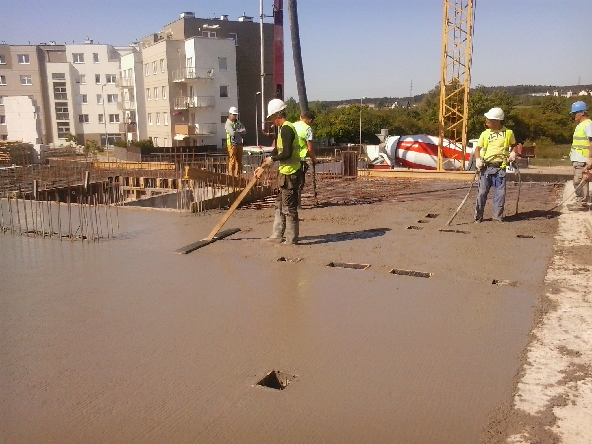 Wylewanie betonu na budowie, widoczni pracownicy w kaskach i kamizelkach odblaskowych, w tle budynki mieszkalne i żuraw budowlany.