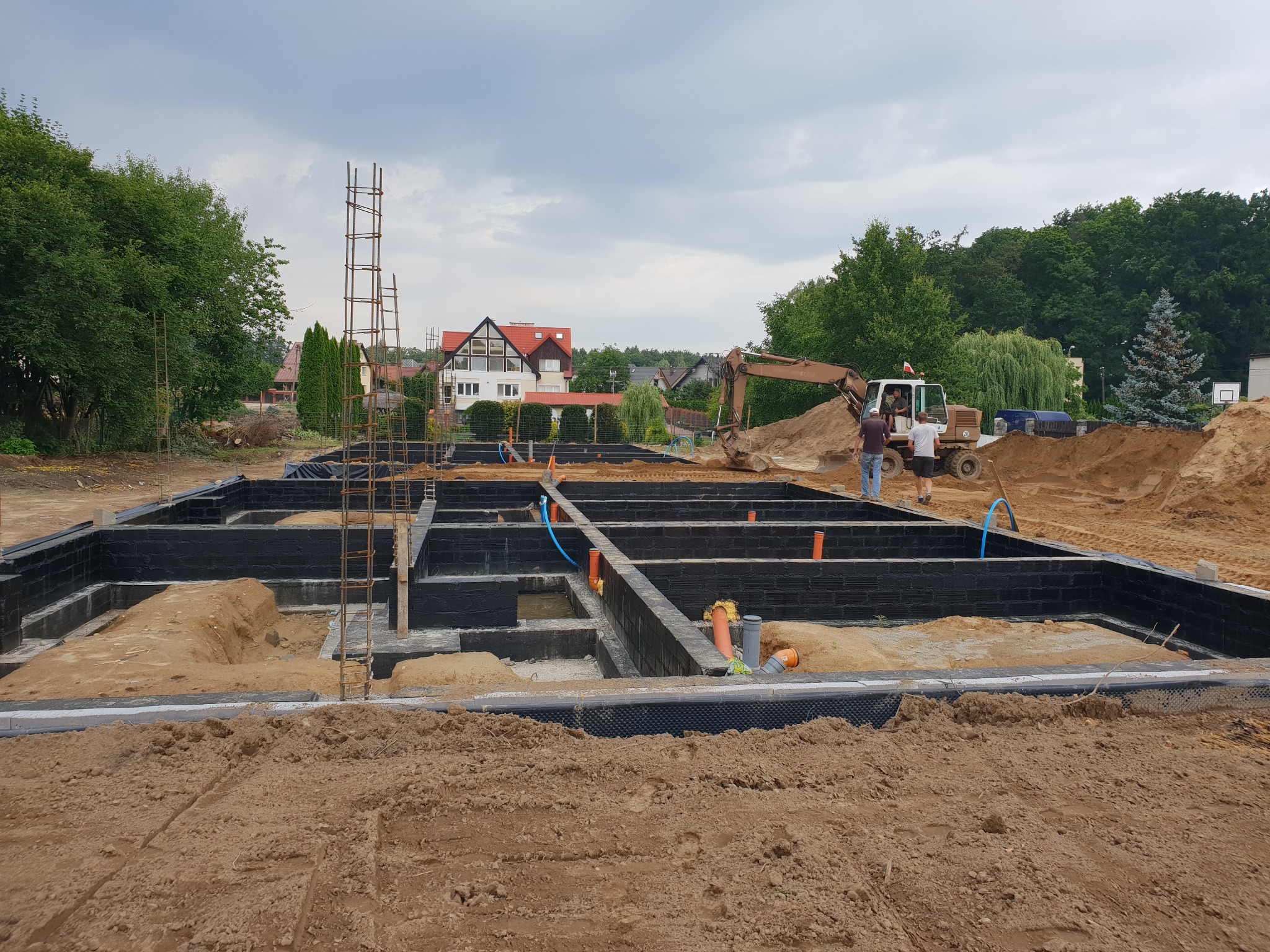 Wykopane fundamenty domu jednorodzinnego z hydroizolacją, zbrojeniami i instalacjami, z widoczną koparką w tle i dwoma pracownikami.